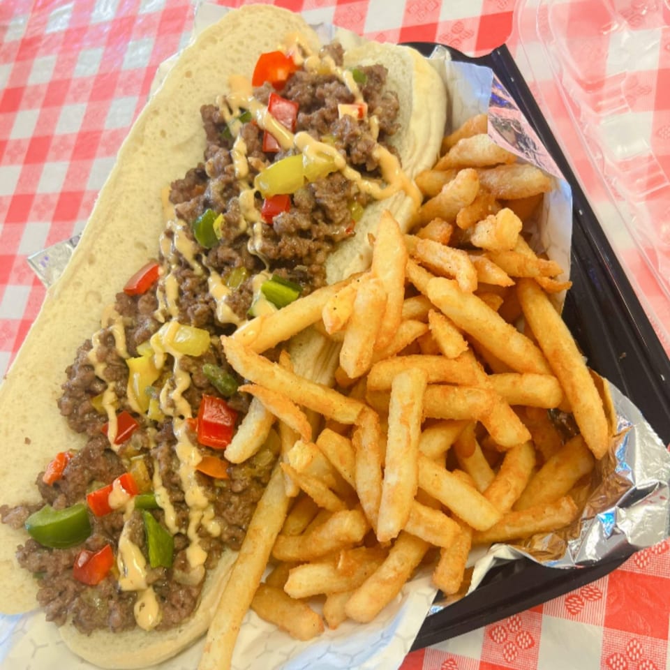 Classic Gourmet Deli's cheesesteak with peppers and sauce, served with golden french fries on a red checkered tablecloth.