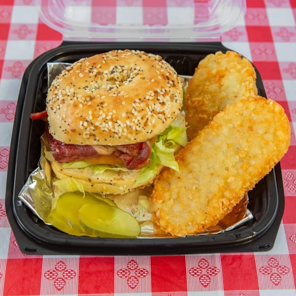 Classic Gourmet Deli bagel sandwich with bacon, lettuce, tomato, and hash browns on a red checkered tablecloth.
